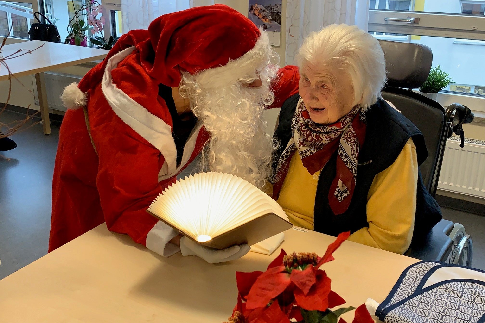 Weihnachtsmann liest Seniorin im Rollstuhl ein Buch vor.