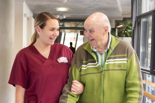 Pflegekraft in roter Uniform lächelt mit älterem Mann im Krankenhausflur.