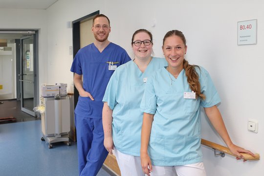Drei Pflegekräfte in Krankenhausflur, lächelnd und in blauer Arbeitskleidung.