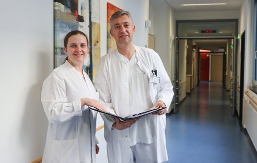 Zwei Ärzt:innen in weißen Kitteln stehen im Klinikflur und lächeln.