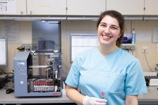 Lächelnde Laborantin in blauer Uniform vor medizinischem Analysegerät im Labor.