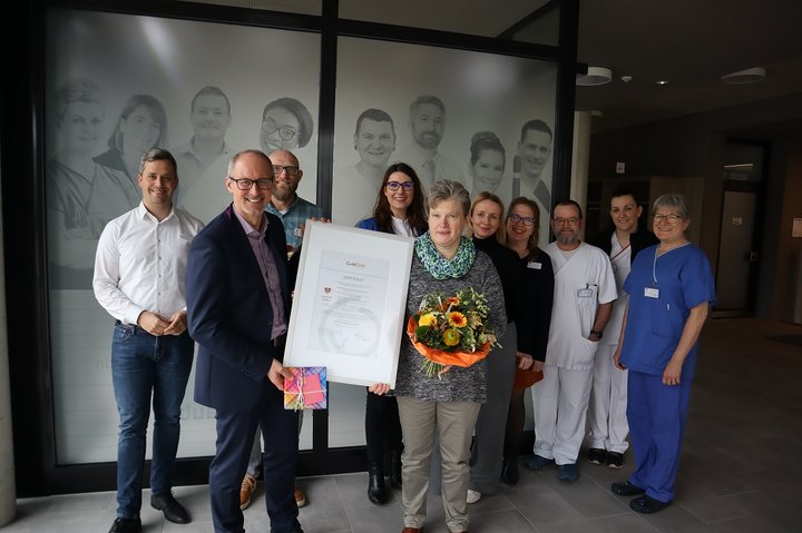 Team erhält Zertifikat und Blumenstrauß vor Klinik-Hintergrund.