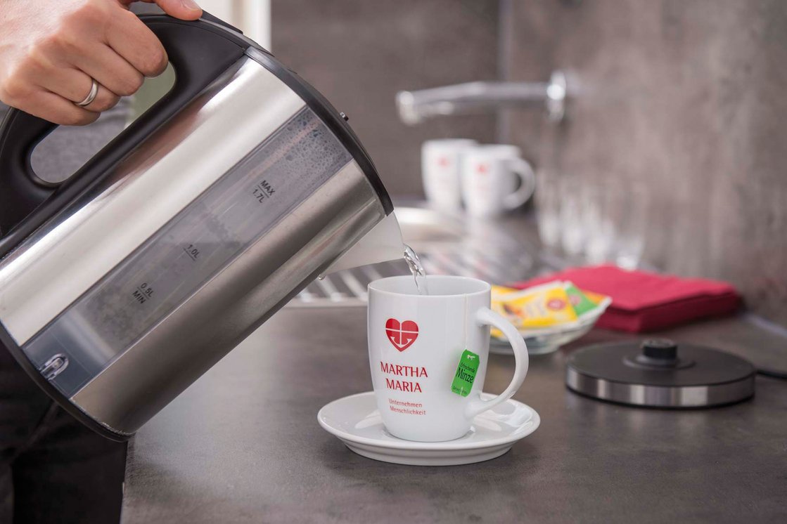 Heißwasserkocher füllt Tasse mit Martha-Maria Logo in einer Küche.