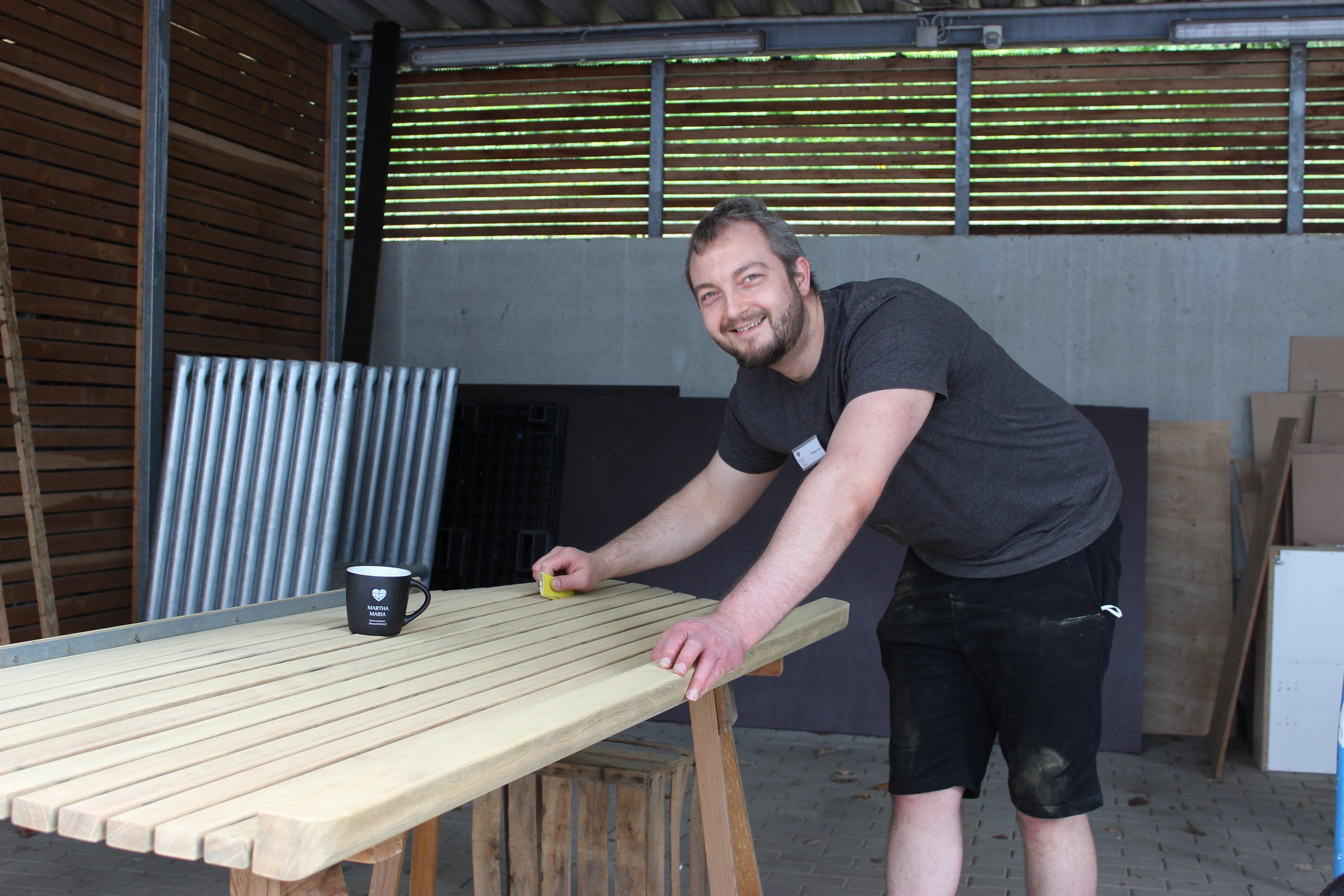 Mann schleift Holztisch in Werkstatt, lächelnd und mit Kaffeetasse.