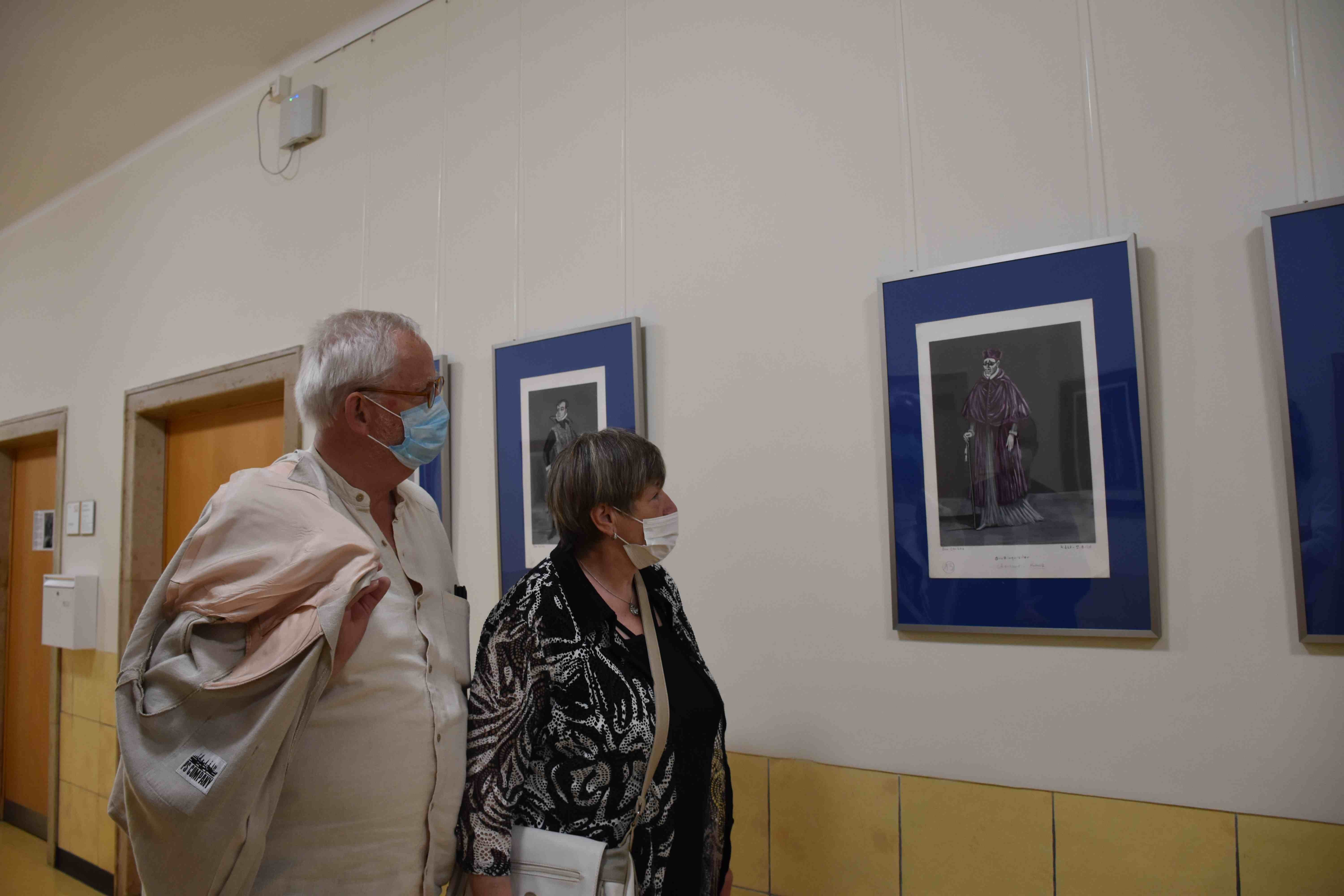 Zwei Personen mit Masken betrachten Kunstwerke an einer Galeriewand.