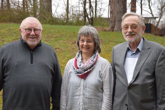 Drei Menschen stehen lächelnd im Freien vor einer grünen Wiese.