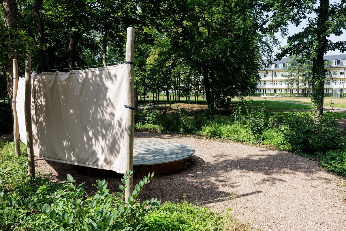 Holzplattform mit Stoffbahnen im besinnlichen Krankenhausgarten umgeben von Bäumen.