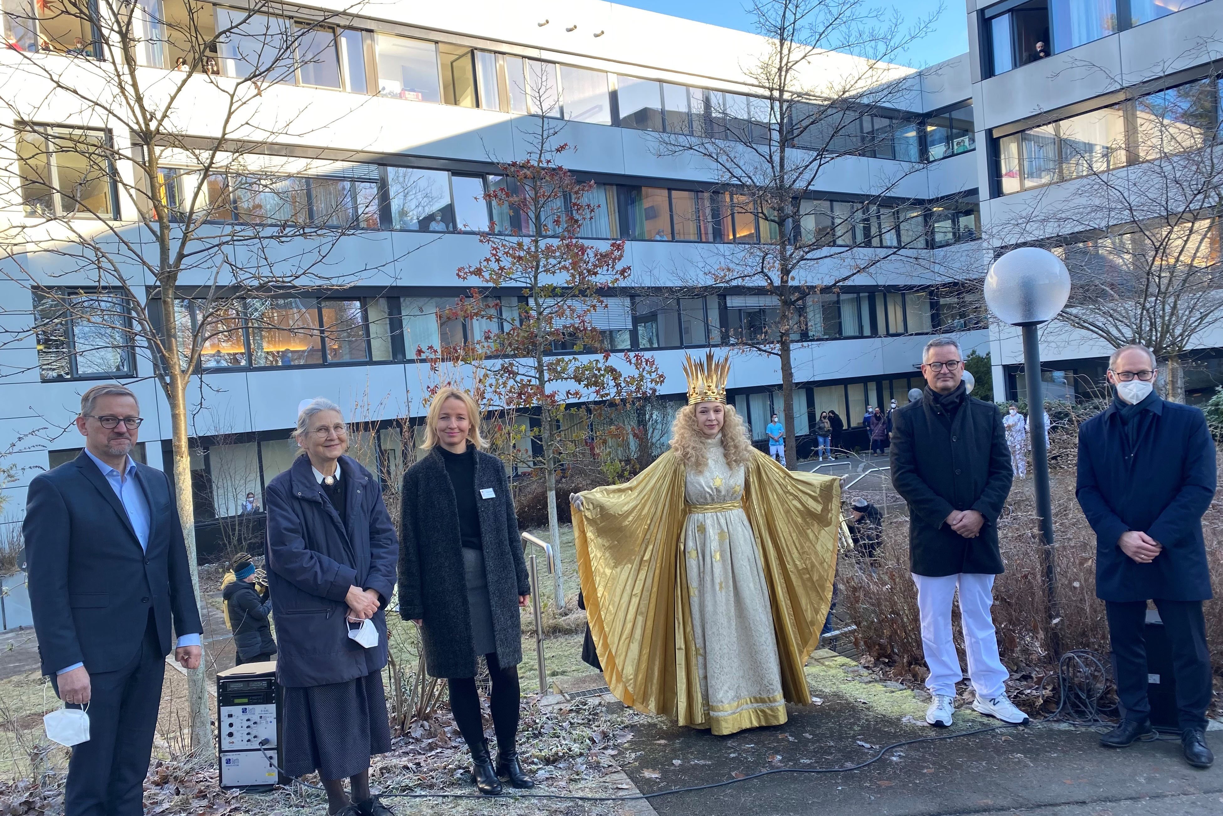 Gruppe von Menschen im Freien vor einem modernen Gebäude mit Christkind.