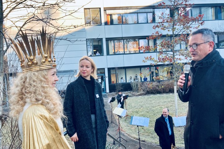 Drei Personen im Freien vor einem Gebäude, eine mit goldener Krone.