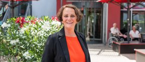 Lächelnde Frau in orangem Shirt vor Pflegeheim mit Sonnenschirmen und Blumen.