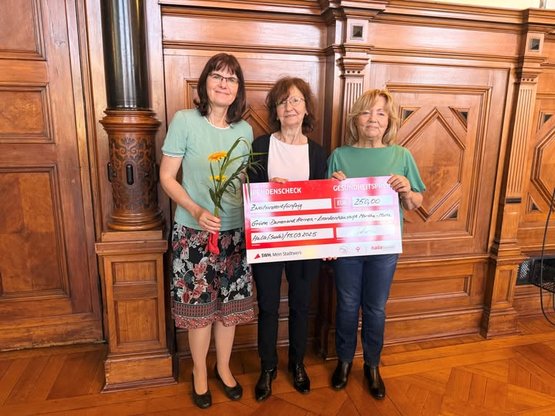 Drei Frauen halten einen Scheck für 250 Euro in einem holzgetäfelten Raum.