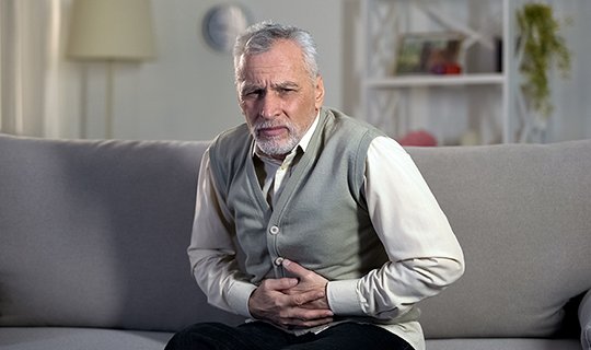 Älterer Mann mit Bauchschmerzen sitzt auf Sofa, hält sich den Bauch.