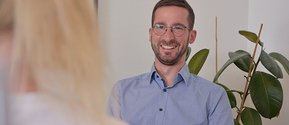 Lächelnder Mann im Gespräch mit einer anderen Person, Pflanze im Hintergrund.
