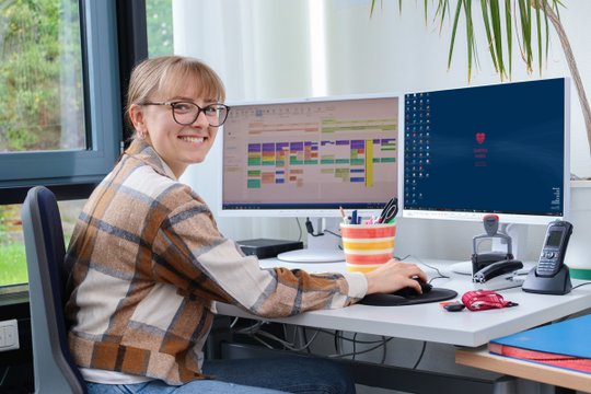Lächelnde Frau am Schreibtisch, arbeitet am Computer mit zwei Bildschirmen.