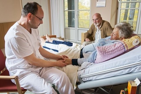 Arzt hält die Hand einer liegenden Patientin, älterer Mann beobachtet.