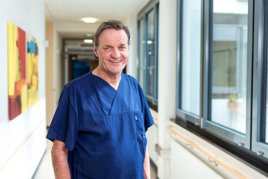 Arzt in blauer OP-Kleidung lächelt im Krankenhausflur der Klinik.