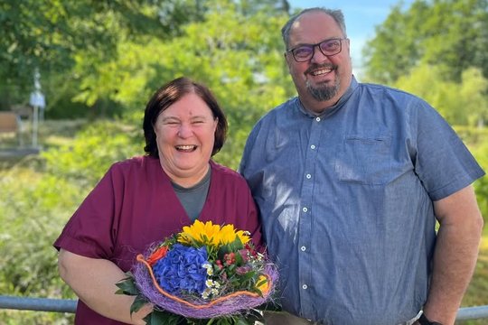 Lächelnde Frau mit Blumenstrauß, Mann daneben, beide draußen.
