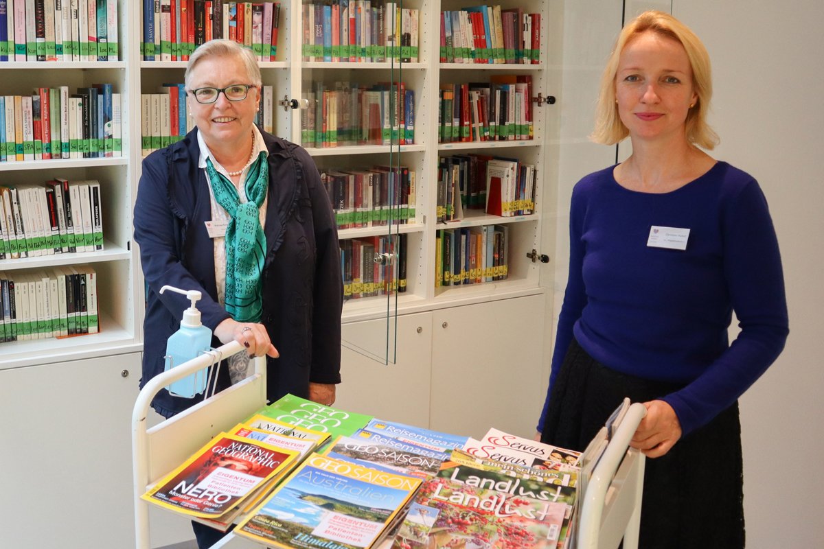 Zwei Frauen in einer Bibliothek mit Bücherregalen im Hintergrund.