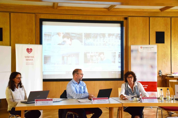 Drei Personen diskutieren vor einem Projektorbild in einem Konferenzraum.