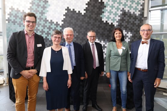 Sechs Personen stehen lächelnd vor einer Wand aus Mosaikmustern.