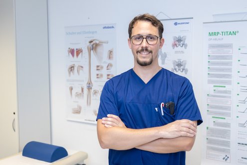 Arzt in blauer Uniform steht lächelnd vor medizinischen Postern in einem Behandlungsraum.