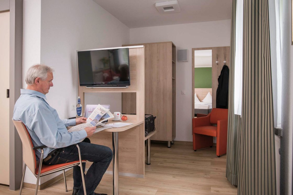 Älterer Mann liest Zeitung in gemütlichem Gästezimmer mit Fernseher und Schreibtisch.