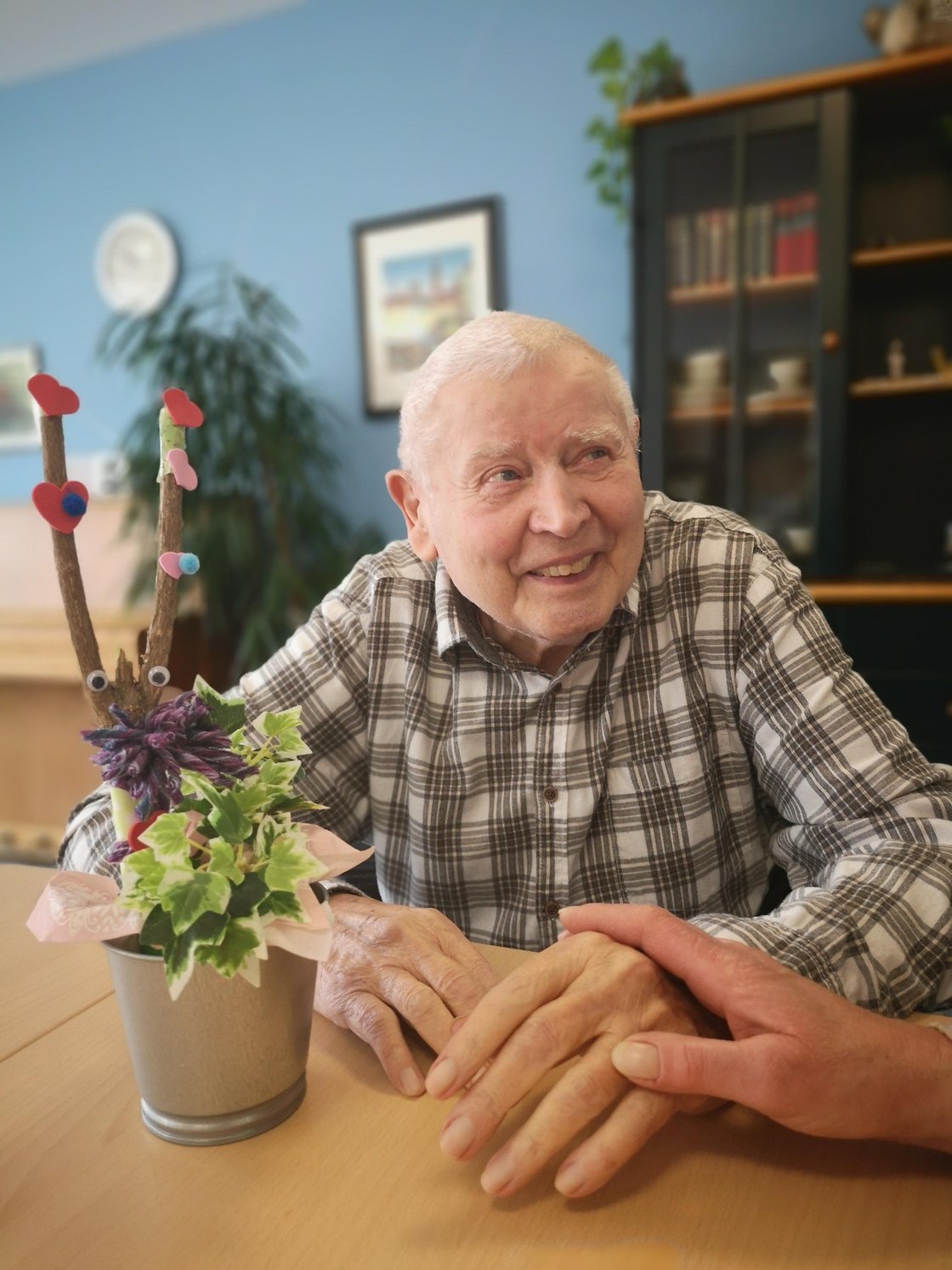 Älterer Mann lächelt am Tisch mit Pflanzenarrangement, Hand gehalten.