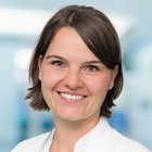 Lächelnde Frau in weißem Kittel vor unscharfem Hintergrund.