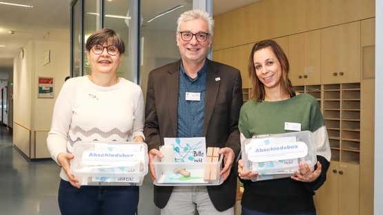Drei Personen halten Abschiedsboxen in einem Krankenhausflur, symbolisiert würdevoller Abschied.