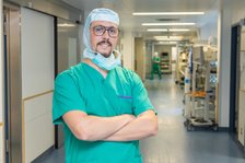 Arzt im grünen Kittel und Haube steht mit verschränkten Armen im Krankenhausflur.