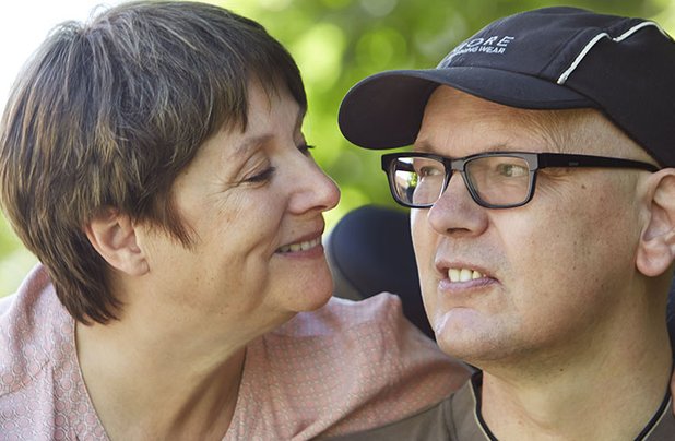 Frau lächelt Mann im Rollstuhl im Freien liebevoll an.