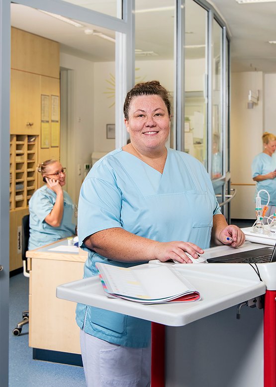 Pflegekraft in Krankenhausumgebung, lächelnd, mit Computer und Dokumenten.