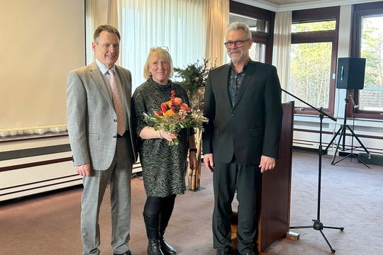 Drei Personen, eine Frau mit Blumenstrauß, posieren lächelnd im Saal.