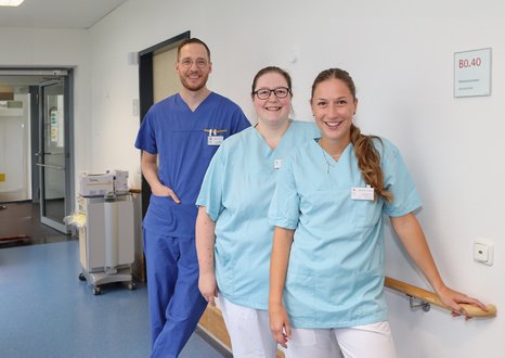 Drei Pflegekräfte in Krankenhausflur, lächelnd und in blauer Arbeitskleidung.