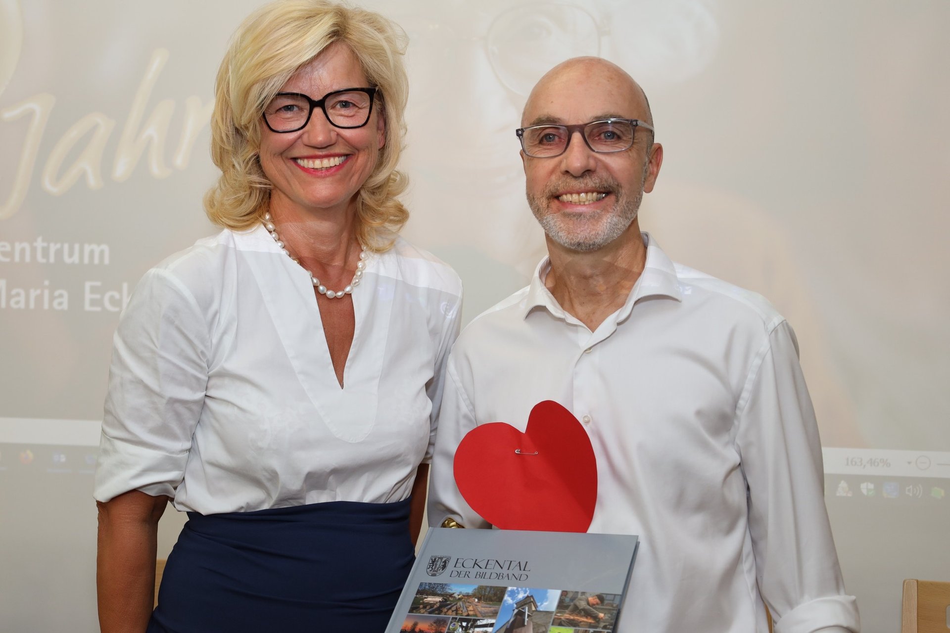 Zwei lächelnde Personen mit Brille, eine hält ein rotes Herz und ein Buch.