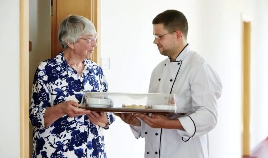 Koch überreicht Tablett mit Essen an ältere Frau im Krankenhausflur.