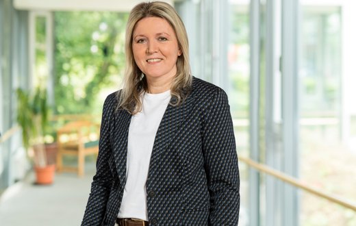 Renata Petrova lächelt in einem sonnigen Flur mit Pflanzen im Hintergrund.