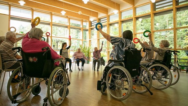 Senioren im Rollstuhl machen Gymnastikübungen mit bunten Reifen in einem Gemeinschaftsraum.